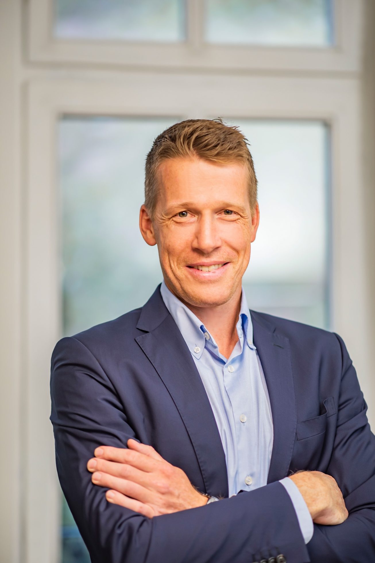 Männlicher Geschäftsmann in Anzug lächelt vor einem hellen Fenster.